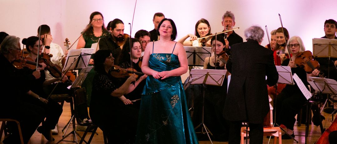 Eine Frau in einem blauen Kleid steht auf der Bühne und schaut in Richtung des Orchesters. Das Orchester mit Violinen und Celli steht hinter ihr. Der Dirigent, ein älterer Mann, befindet sich auf der rechten Seite der Bühne.