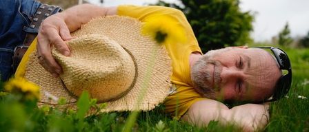 Ein Mann in einem gelben Shirt und einem Strohhut liegt auf dem Gras, die Augen geschlossen, und trägt eine Sonnenbrille.