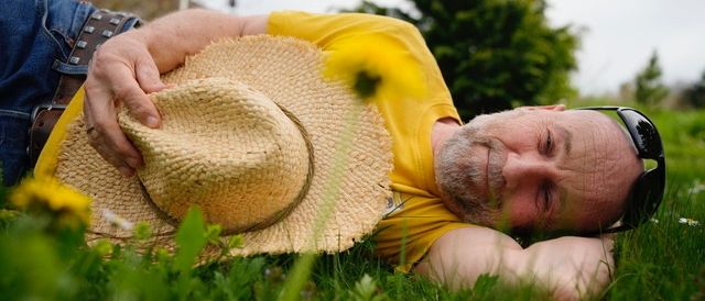Ein Mann in einem gelben Shirt und einem Strohhut liegt auf dem Gras, die Augen geschlossen, und trägt eine Sonnenbrille.