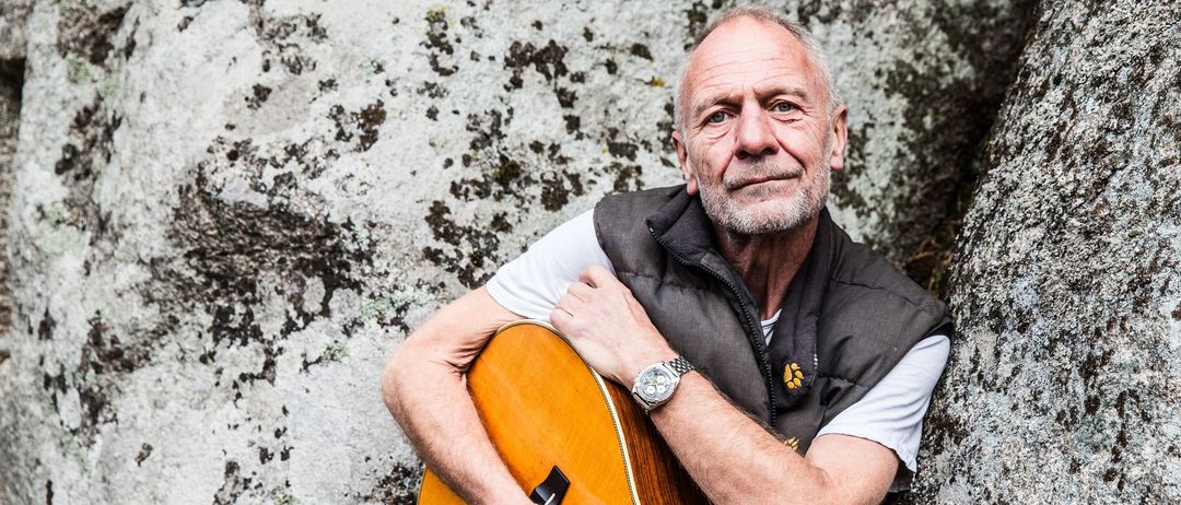 Ein älterer Mann mit einer Gitarre lehnt sich gegen einen Felsen. Er hat eine Uhr am linken Handgelenk und ein leichtes Lächeln.