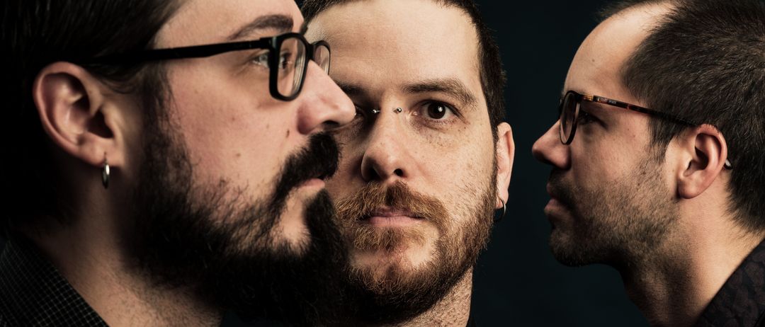 Drei Männer mit Bärten und Brillen schauen sich gegenseitig an, einer mit einem Piercing, vor dunklem Hintergrund.