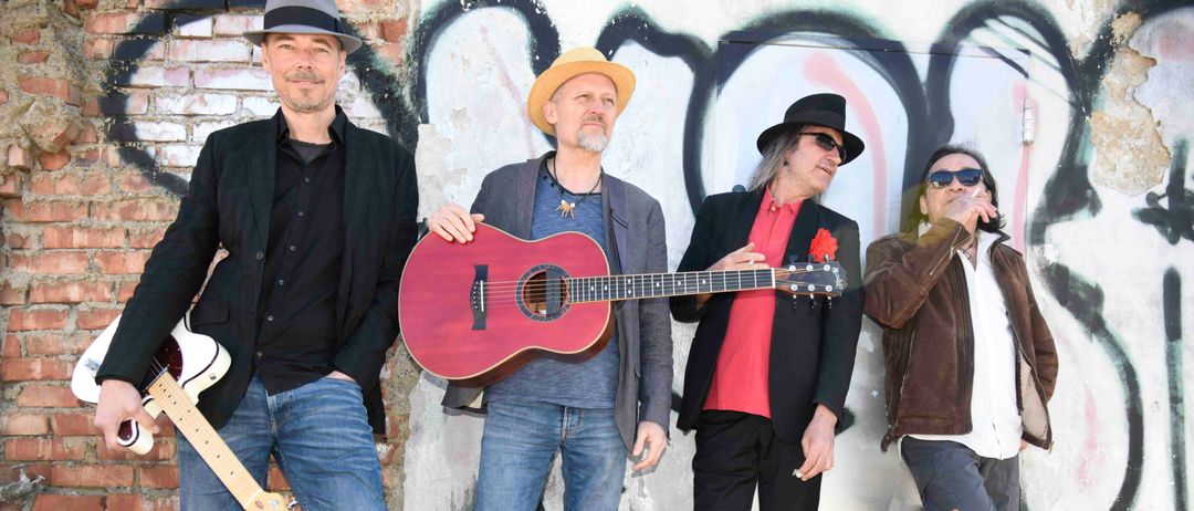 Vier Männer stehen vor einer mit Graffiti bedeckten Wand. Der mittlere Mann hält eine rote Gitarre, während die anderen Gitarren in den Händen oder Taschen haben. Sie tragen Hüte, Sonnenbrillen und lässige Kleidung.