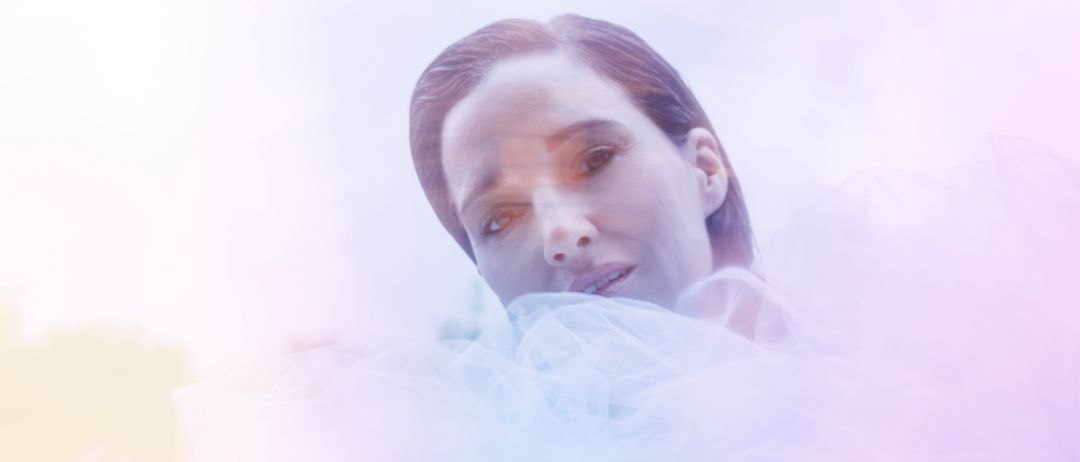 Ein Nahaufnahme des Gesichts einer Frau mit kurzen Haaren, umgeben von weichem, hellfarbenem Stoff, möglicherweise ein Schleier. Sie lächelt leicht und ihre Augen sind leicht geschlossen. Der Hintergrund ist unscharf.