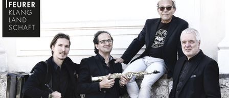 Vier Männer posieren für ein Foto. Der Mann auf der linken Seite hält einen Saxophonkoffer. Der Mann in der Mitte hält ein Saxophon. Der Mann auf der rechten Seite hält ein rundes Instrument. Sie sind alle in Schwarz gekleidet. Der Hintergrund ist eine Steinmauer.