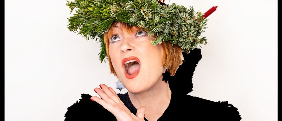 Eine Frau trägt einen schwarzen Pullover mit einem Rentieraufdruck und eine Krone aus Tannenzweigen mit brennenden Kerzen auf dem Kopf.