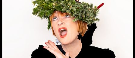 Eine Frau trägt einen schwarzen Pullover mit einem Rentieraufdruck und eine Krone aus Tannenzweigen mit brennenden Kerzen auf dem Kopf.