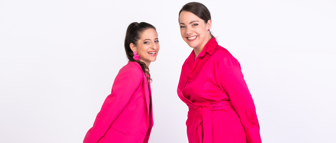 Zwei Frauen in leuchtend rosa Outfits, eine mit Blazer und die andere mit Kleid, stehen eng beieinander und lächeln.