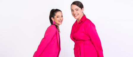 Zwei Frauen in leuchtend rosa Outfits, eine mit Blazer und die andere mit Kleid, stehen eng beieinander und lächeln.