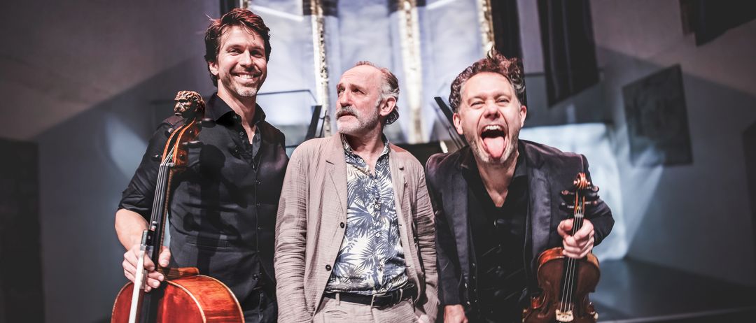 Drei Männer in einem Studio, einer mit einer Geige, ein anderer mit einem Cello und einer mit einem lächelnden Gesicht. Sie stehen zusammen und scheinen für ein Foto zu posieren.