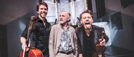 Drei Männer in einem Studio, einer mit einer Geige, ein anderer mit einem Cello und einer mit einem lächelnden Gesicht. Sie stehen zusammen und scheinen für ein Foto zu posieren.