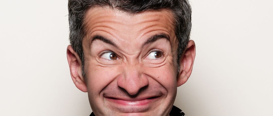 Ein lächelnder Mann mit kurzen Haaren, der ein schwarzes Blumenhemd und eine Halskette trägt, posiert für ein Bild.