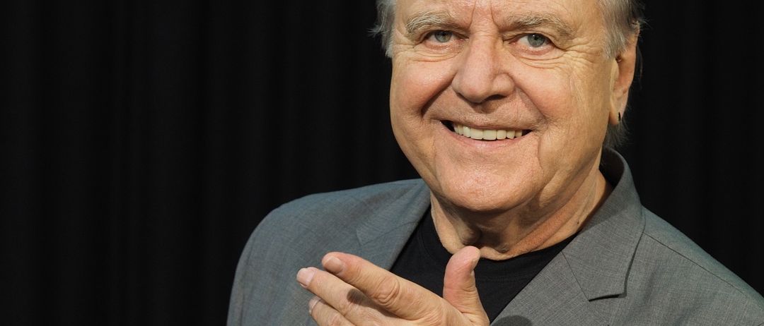 Ein älterer Mann mit grauem Haar, der einen grauen Anzug und ein schwarzes Shirt trägt, lächelt und gibt einen Daumen hoch. Er trägt auch eine silberne Uhr am linken Handgelenk.