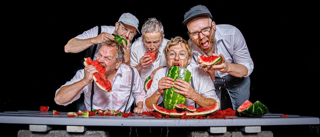 Fünf Männer essen Wassermelonen auf einer Bank und machen einen Schlamassel. Sie tragen alle weiße Hemden und Hosenträger. Zwei von ihnen tragen Hüte. Die Szene ist vor einem dunklen Hintergrund.