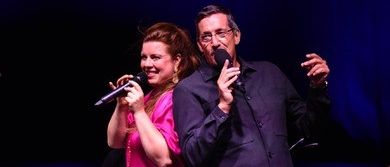 Ein Mann und eine Frau stehen auf der Bühne, halten Mikrofone und singen, während eine Band im Hintergrund spielt.