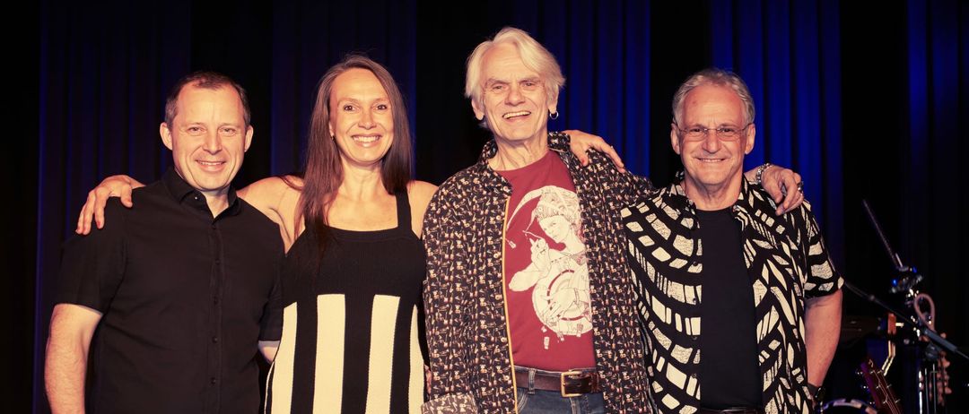 Vier Personen posieren für ein Foto auf einer Bühne mit blauen Vorhängen. Der Mann links trägt ein schwarzes Hemd und Jeans, während die anderen gemusterte Hemden und Jeans tragen.