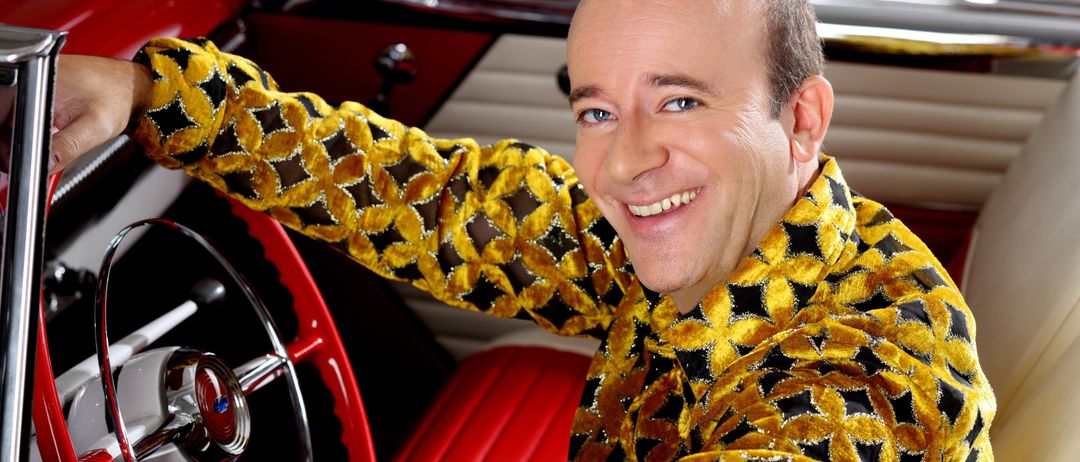 Ein lächelnder Mann in einem gelben und schwarzen gemusterten Hemd sitzt auf dem Fahrersitz eines klassischen roten Autos. Er hält das Lenkrad mit einer Hand und sein Lächeln ist strahlend.