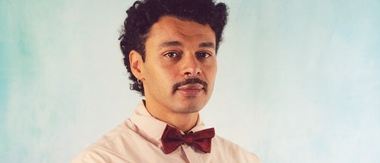 Ein Mann mit lockigem Haar und einem Schnurrbart steht vor einer blauen Wand. Er trägt ein rosa Hemd mit einer Fliege.