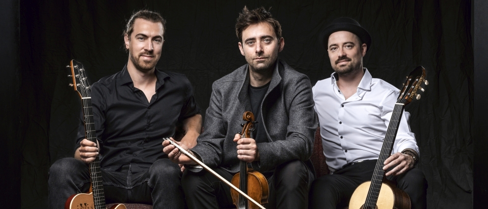 Drei Männer sitzen gemeinsam auf einer Couch, jeder hält ein Musikinstrument in der Hand, eines eine Gitarre, ein anderes eine Geige und der dritte eine Gitarre. Der Mann rechts trägt einen Hut und der Mann links hat einen Bart.