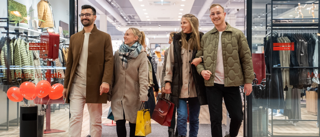 Vier junge Menschen betreten einen Bekleidungsladen, lächeln und halten Taschen. Ein Mann links trägt eine Brille und einen braunen Mantel. Eine Frau neben ihm hat einen Schal und einen hellen Mantel. Eine andere Frau rechts trägt eine schwarze Jacke. Der Mann rechts trägt einen grünen Mantel und eine Brille.