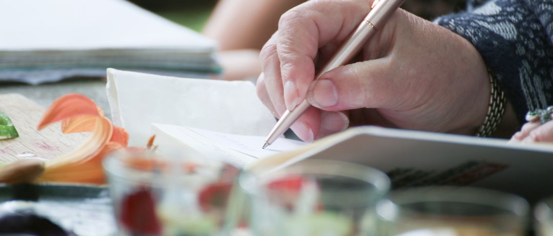 Eine Hand schreibt mit einem Stift auf einem Stück Papier auf einem Tisch mit Essen und Getränken.