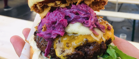 Ein Nahaufnahme eines Gourmet-Burgers mit einem gebratenen Ei, geschmolzenem Käse und lila Kraut, gehalten von einer Hand auf einem Outdoor-Food-Festival.
