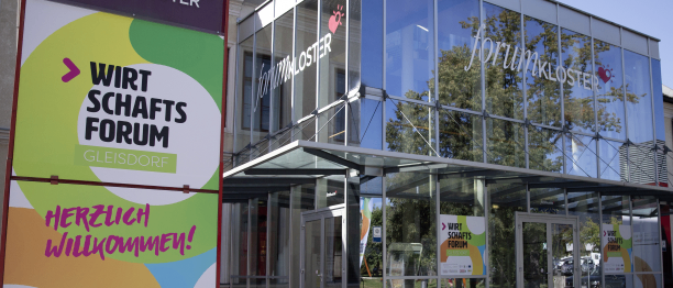 Der Eingang zu einem modernen Gebäude mit Glaswänden und einem Willkommensschild auf Deutsch. Das Gebäude ist von einem gepflasterten Bereich und einem Baum umgeben.