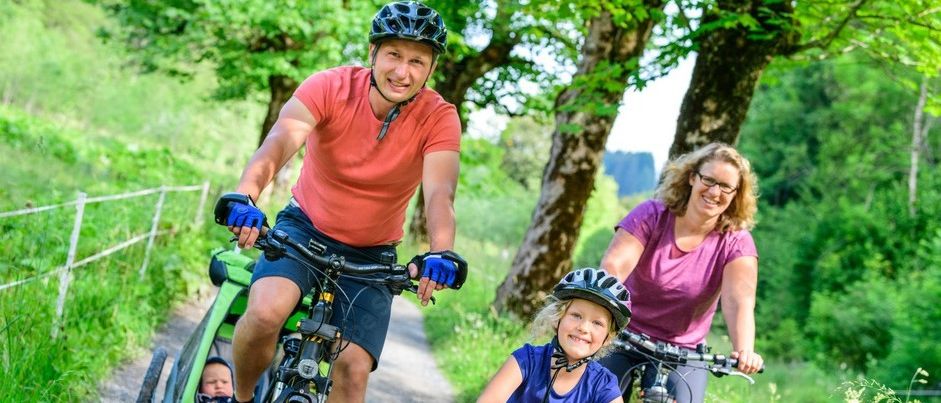 Eine vierköpfige Familie fährt auf einem Kiesweg Rad. Der Vater fährt ein Fahrrad mit Kindersitz, die Mutter fährt ein Fahrrad hinter ihm, und die beiden Kinder fahren ihre eigenen Fahrräder. Sie tragen alle Helme und scheinen die Fahrt zu genießen.