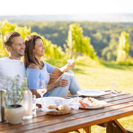 Ein Paar sitzt an einem Tisch in einem Weinberg und lächelt. Sie haben Wein und Essen auf dem Tisch.