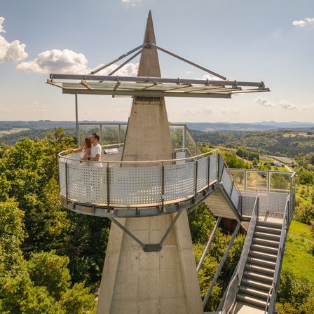 Ein Paar steht auf dem Balkon eines hohen Turms und genießt einen Panoramablick auf die grüne Landschaft unten.
