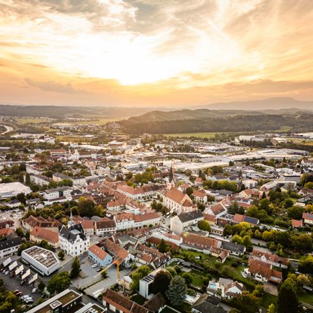Eine Luftaufnahme einer Stadt bei Sonnenuntergang mit Gebäuden verschiedener Größen und Stile, umgeben von Bäumen und Bergen in der Ferne.