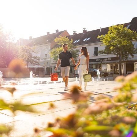 Ein Paar geht Hand in Hand durch einen Stadtplatz mit Springbrunnen und trägt Einkaufstüten. Der Himmel ist klar und sonnig. Dahinter säumen Gebäude die Straße, darunter ein Geschäft namens Anita Tieber.