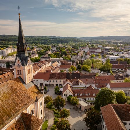 Luftaufnahme einer Stadt mit Gebäuden, Bäumen und einer Kirche mit Turm und Uhr. Berge sind in der Ferne sichtbar.