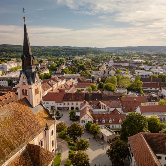 Luftaufnahme einer Stadt mit Gebäuden, Bäumen und einer Kirche mit Turm und Uhr. Berge sind in der Ferne sichtbar.