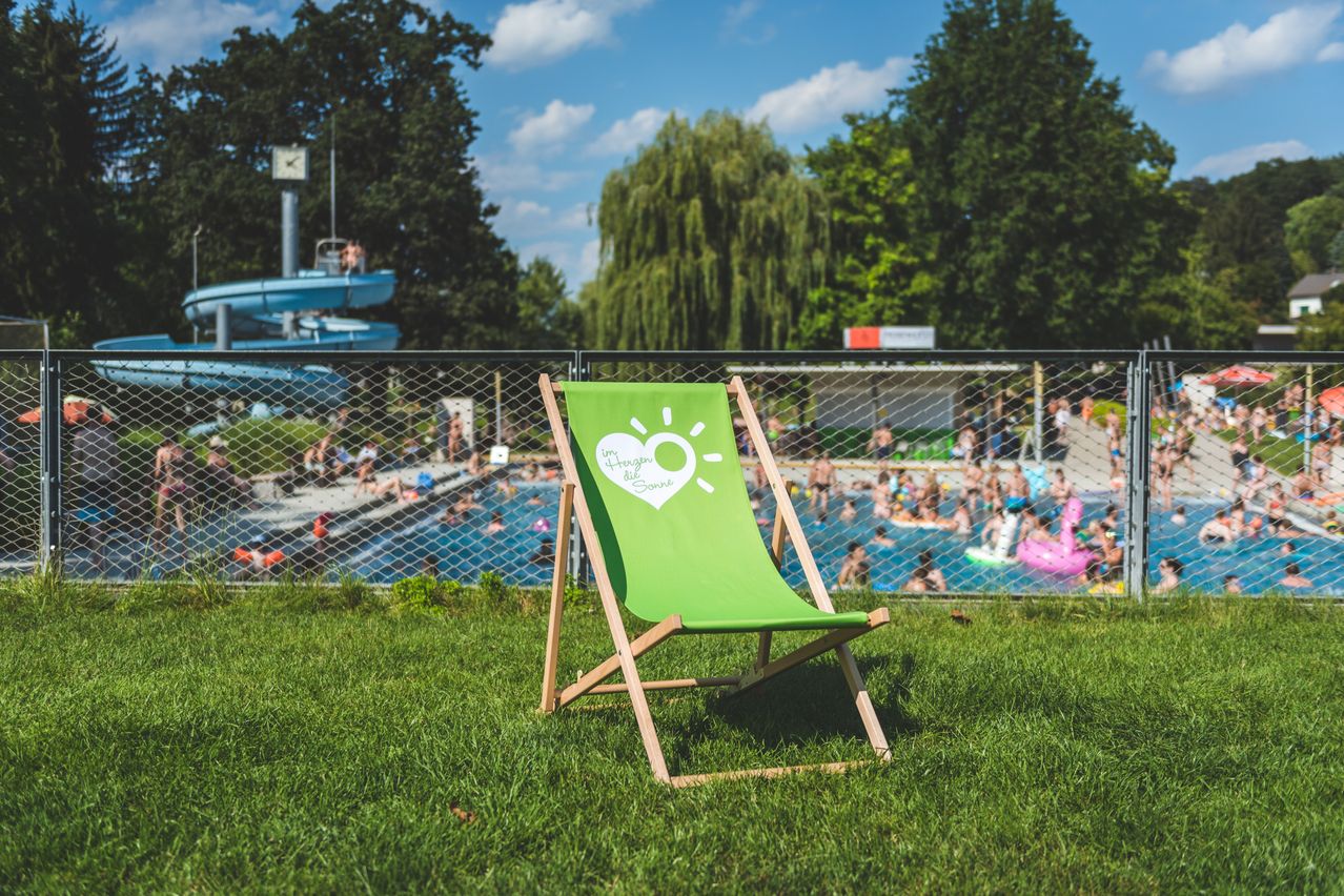 Ein grüner Liegestuhl auf einer Wiese mit einem Pool und Menschen, die im Hintergrund schwimmen.