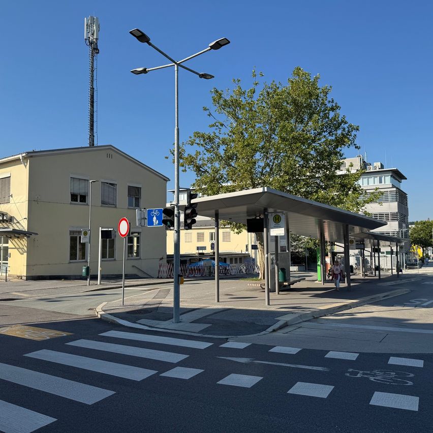 Eine Außenszene mit einer Bushaltestelle unter einem überdachten Dach, einer Verkehrsampel, einem Fahrradschild und einem Baum. Ein Gebäude mit Fenstern und einem Turm steht links.