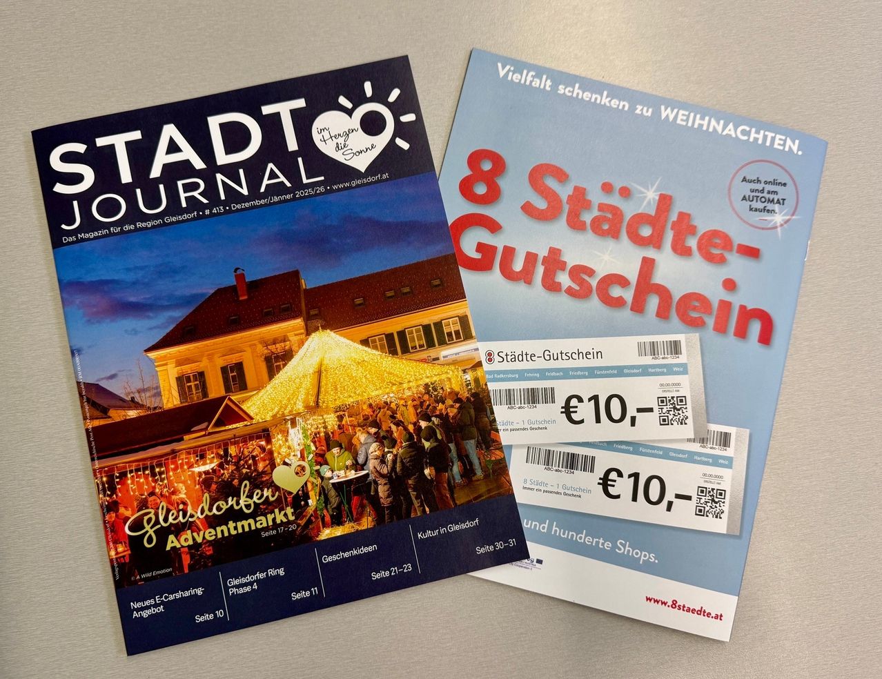 Zwei Broschüren für eine lokale Veranstaltung werden gezeigt. Eine zeigt einen Weihnachtsmarkt mit Menschen, Lichtern und einem Zelt, während die andere ein Geschenkgutschein mit dem Text 'Vielfalt schenken zu WEIHNACHTEN' ist.