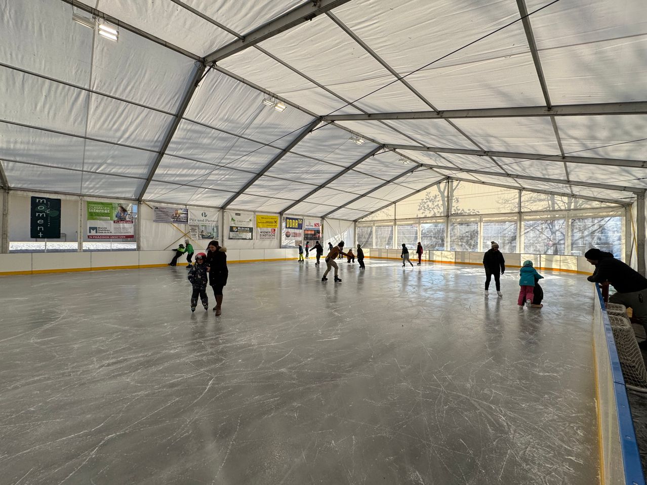 Eine überdachte Eislaufbahn mit weißem Zeltdach, auf der mehrere Kinder und Erwachsene unter weißen Lichtern Schlittschuh laufen. An den Wänden sind Werbetafeln angebracht.