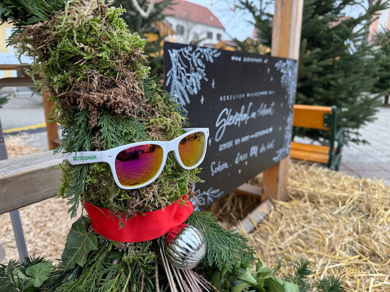 Ein mit einem roten Band und weißen Sonnenbrillen geschmückter Weihnachtsbaum steht vor einem Schild, das 'Schlaflos in Advent' besagt. Dahinter ist eine Holzbank zu sehen.