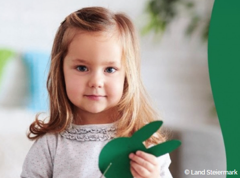 Ein kleines Mädchen mit blonden Haaren, das ein weißes Oberteil trägt, hält einen grünen Papierhasen in den Händen. Sie steht in einem Raum mit einer weißen Wand und verschwommenen grünen Pflanzen.