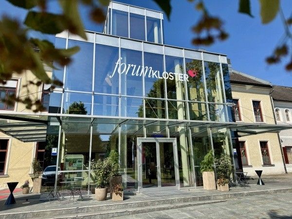 Der moderne Eingang von Forum Kloster mit Glaswänden, einer Glasüberdachung und Topfpflanzen draußen. Ein Auto ist in der Nähe geparkt.