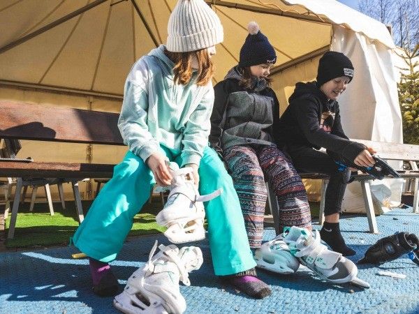 Drei Mädchen sitzen auf einer Bank und stellen ihre Schlittschuhe ein. Sie sind unter einem Zelt, mit Bäumen im Hintergrund.