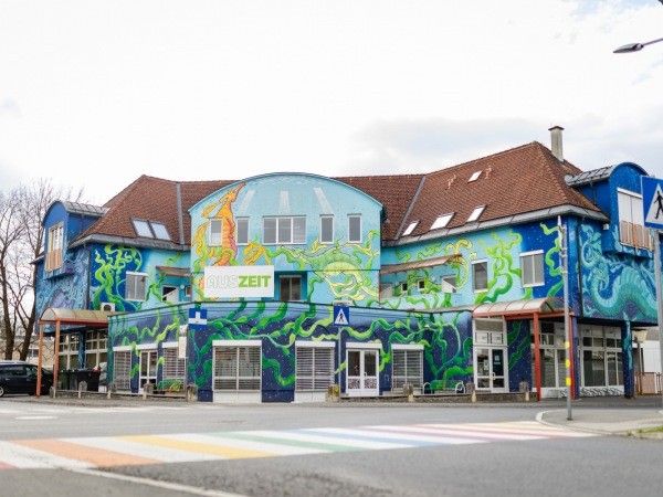 Ein blaues Gebäude mit einem farbenfrohen Wandbild an der Fassade, das verschiedene Kreaturen und Pflanzen zeigt. Es hat ein braunes Dach und mehrere Fenster. Ein Schild mit der Aufschrift 'AUS ZEIT' ist sichtbar.