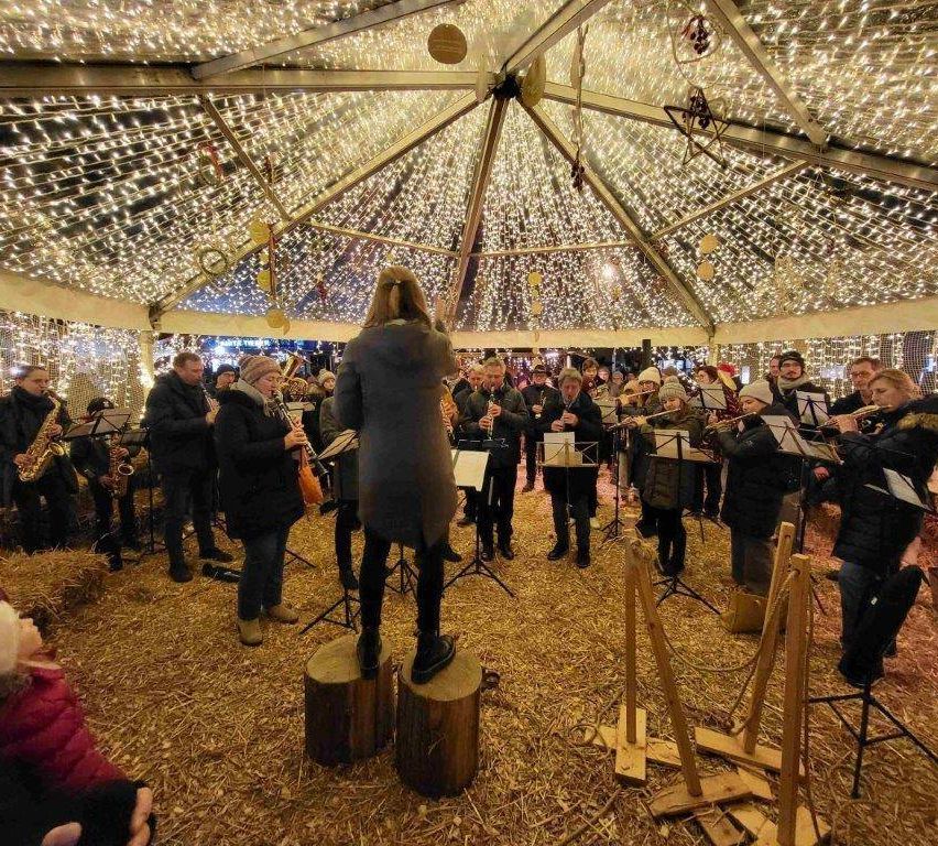 Eine Gruppe von Musikern tritt unter einem beleuchteten Zelt auf. Eine Frau steht auf einem Hocker und leitet die Gruppe. Die Musiker spielen verschiedene Instrumente und das Publikum steht um sie herum.