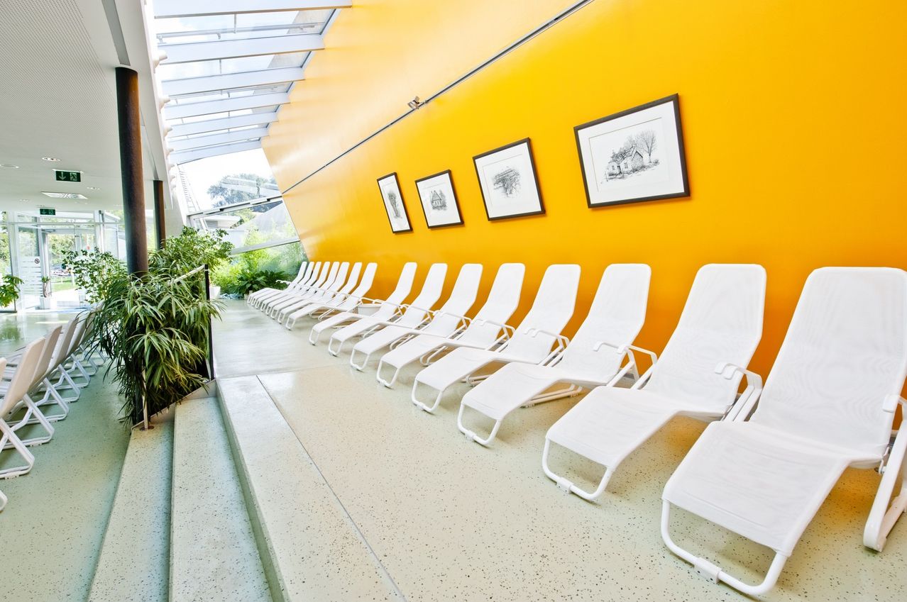 Ein Flur mit gelben Wänden und weißem Boden verfügt über eine Reihe weißer Liegestühle, drei gerahmte Bilder und Grünpflanzen auf der linken Seite.