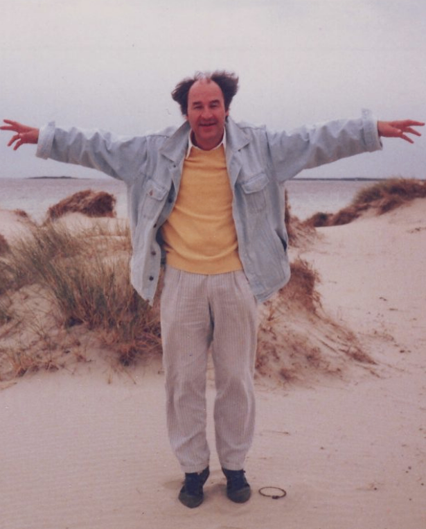 Ein Mann steht auf einem sandigen Strand mit weit ausgestreckten Armen, lächelt und schaut in die Kamera. Hinter ihm befindet sich ein Grasbereich und ein Gewässer.