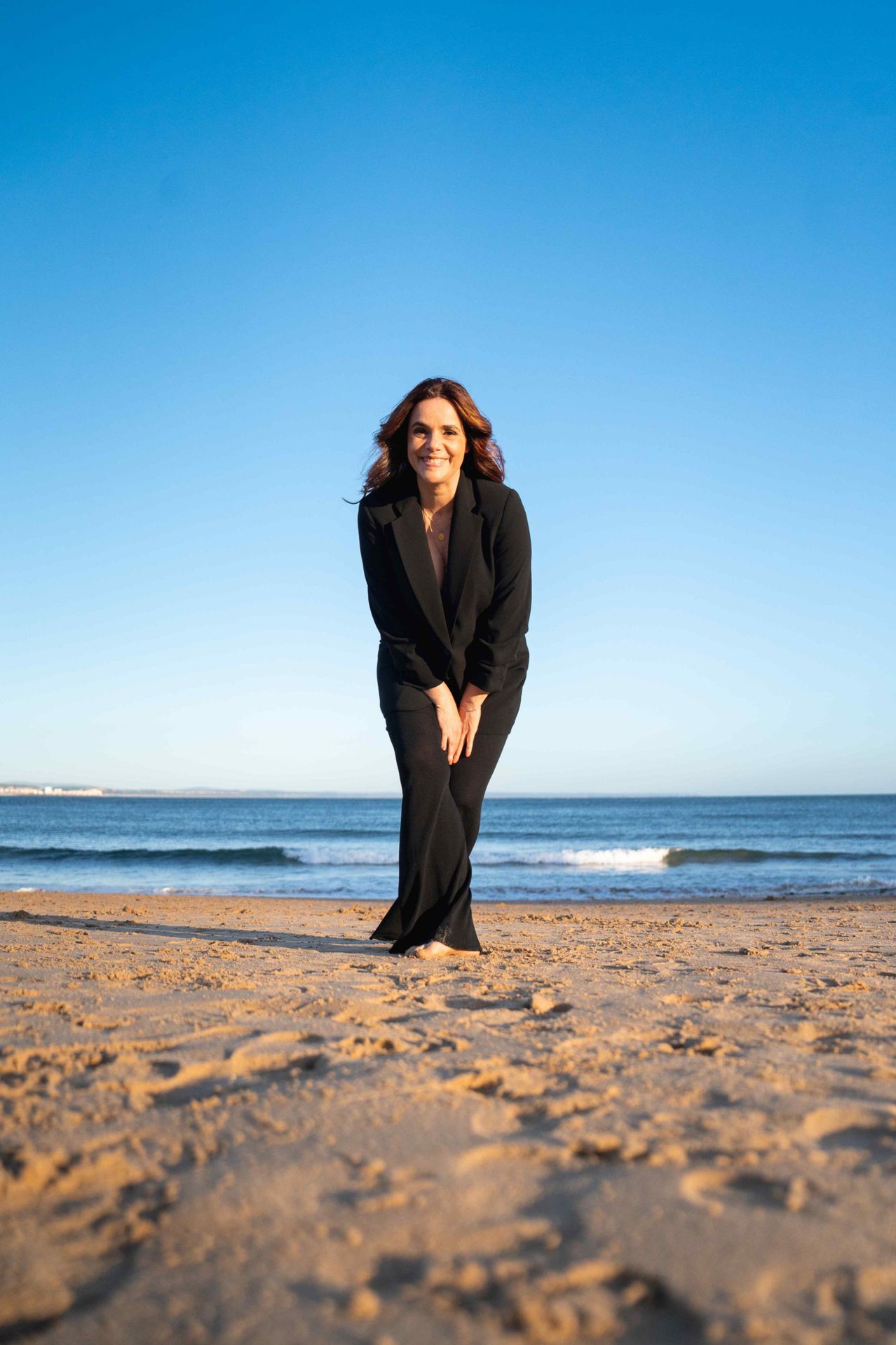 Eine Frau in einem schwarzen Anzug steht am Strand mit einem klaren blauen Himmel und Ozean im Hintergrund.