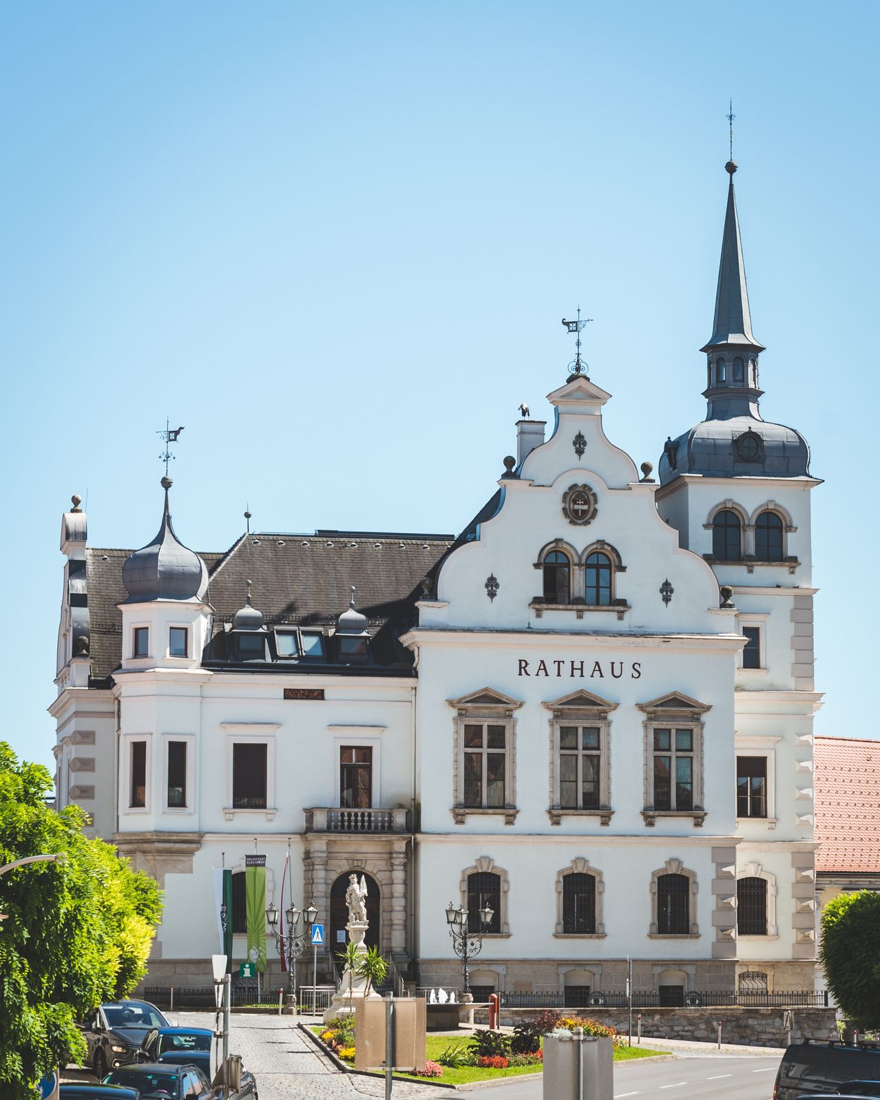 Ein weißes architektonisches Gebäude mit einem spitzen Turm. Es hat ein Namensschild, das Rathaus liest. Es ist von grünen Bäumen umgeben.