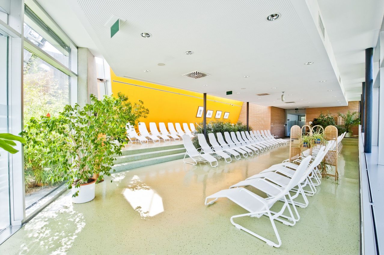 Ein Loungebereich mit in Reihen angeordneten weißen Liegestühlen, einer gelben Wand mit gerahmten Bildern, einer grünen Pflanze in einem Topf und einem glänzenden Boden.