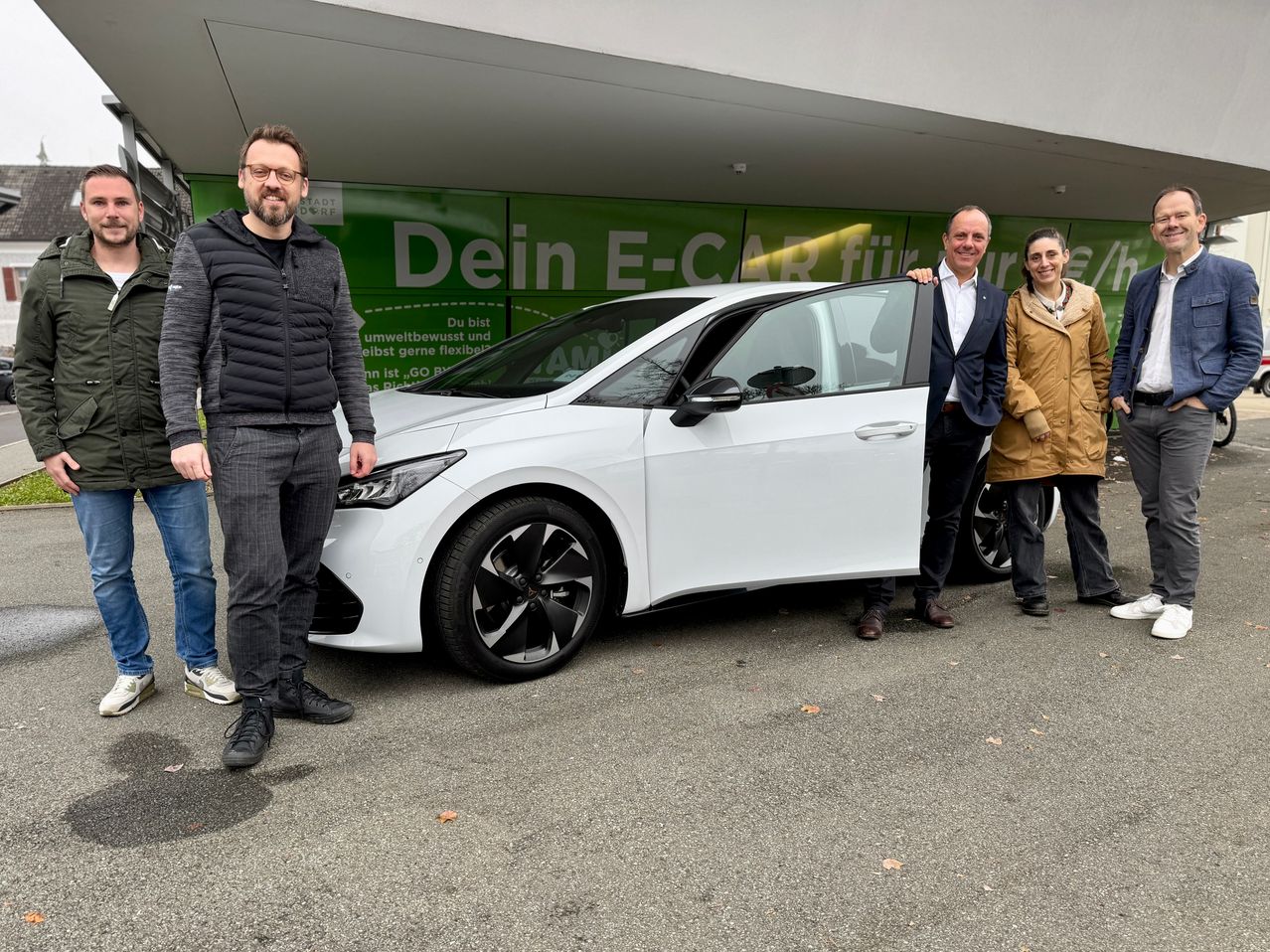 Drei Personen stehen neben einem weißen Elektroauto. Der Mann auf der linken Seite trägt eine schwarze Jacke und eine Brille. Der Mann in der Mitte hält die Autotür. Dahinter steht ein grünes Schild mit der Aufschrift 'Dein E-Car'.