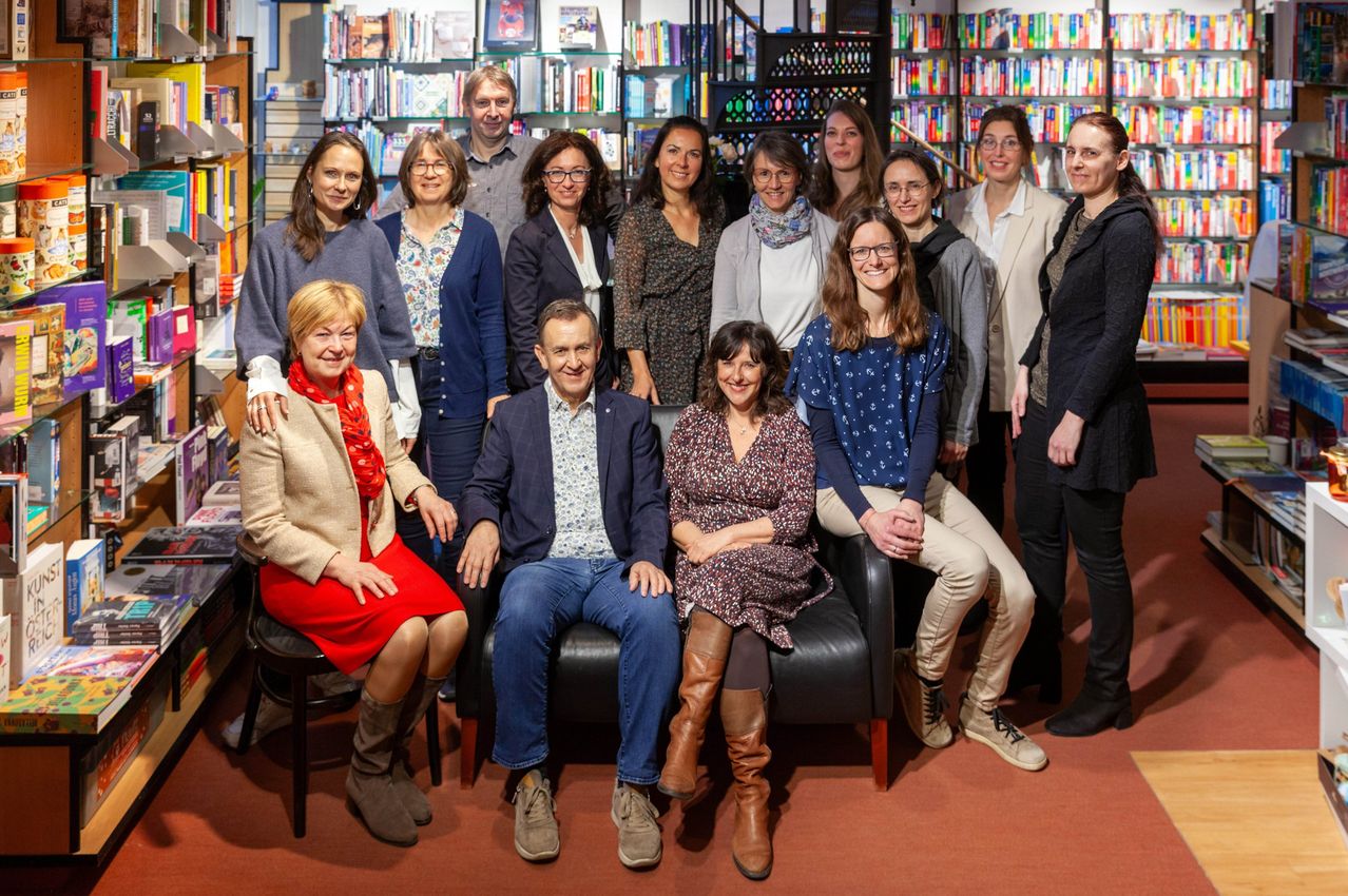 Eine Gruppe von Menschen in einer Bibliothek mit Büchern an den Regalen. Einige Leute stehen, andere sitzen auf Stühlen. Sie tragen alle eine Brille.